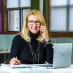 Smiling businesswoman on phone call, working on laptop in sleek office setting.
